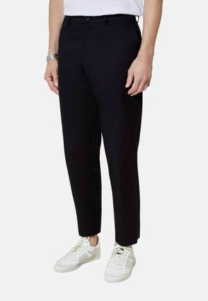 Mężczyzna w czarnych dopasowanych spodniach i białych trampkach, stojący na jednolitym jasnym tle.