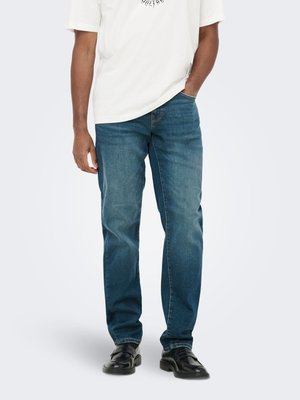 Mężczyzna ubrany w niebieskie dżinsy o prostych nogawkach, czarne mokasyny i białą koszulkę, jedną ręką włożoną do kieszeni.