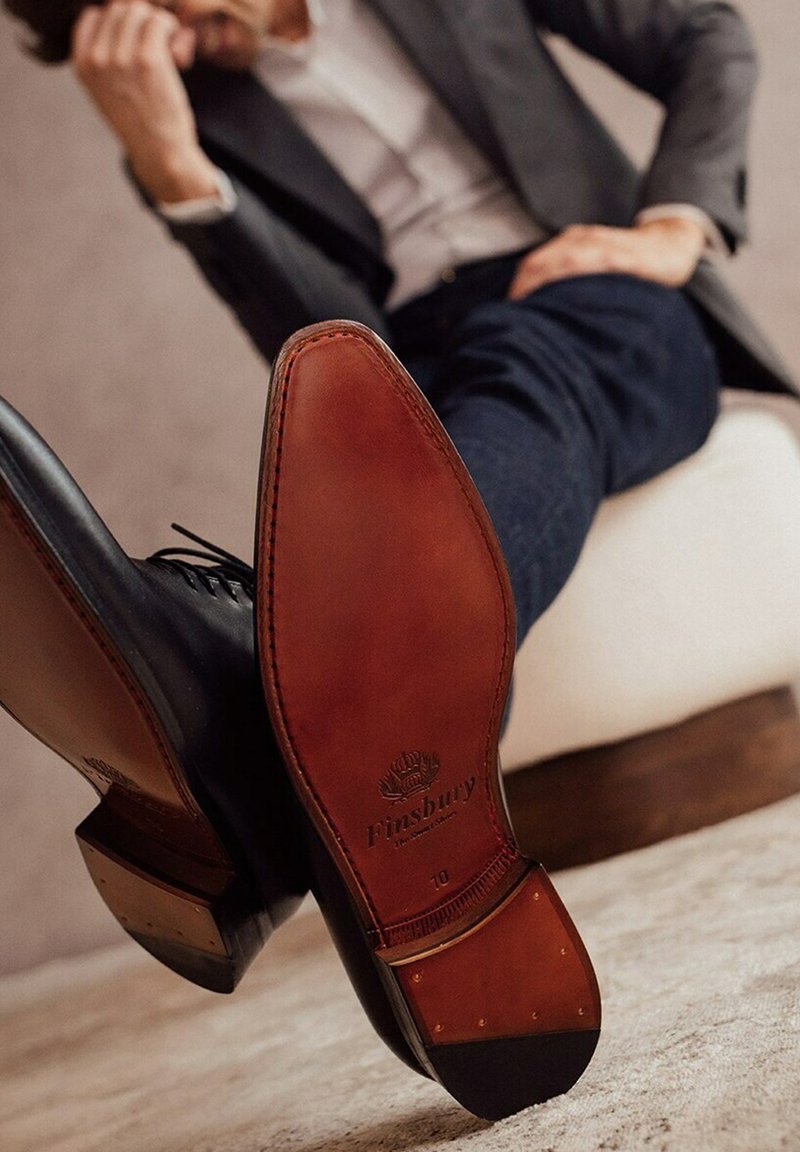 Zwarte leren veterschoen met een slank ontwerp, voorzien van een bruine leren zool, een gegraveerd logo en een metalen accent nabij de hak.