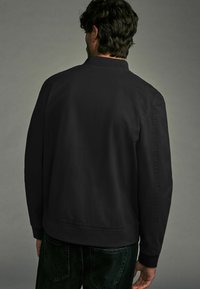 Schwarze Jacke mit geripptem Kragen, glatter Textur, lässiger Passform und elastischem Bund; getragen über dunkelgrünen Hosen, von hinten betrachtet.