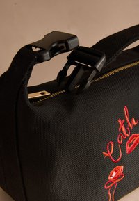 Svart texturerad tygväska med guldfärgad dragkedja, svart plastspänne och det röda broderade ordet "Cathy" med blommönster och läppar.