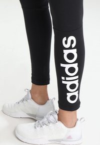 Nohy oblečené v černých legínách Adidas s bílým logem na lýtku a bílých sportovních botách na světlém pozadí.