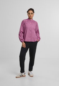 Rosa Hemd mit hohem Kragen und gerafften Details, kombiniert mit schwarzen Hosen und weißen Sneakers mit schwarzem Akzent. Transparente Brille.