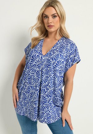 Femme blonde portant un chemisier ample à manches courtes bleu et blanc à motifs avec un col en V et un jean bleu, debout devant un fond uni.