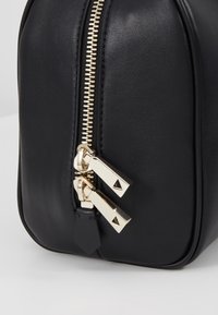 Gros plan sur un coin de sac en cuir noir avec une fermeture éclair dorée et deux curseurs dorés sur un fond clair.