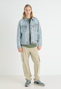 Chaqueta de mezclilla azul claro con bolsillos en el pecho, camisa verde oliva debajo, pantalones cargo beiges con bolsillos laterales y zapatillas grises.