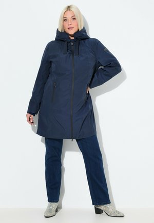 Donna che indossa un cappotto con cappuccio blu navy con zip, jeans blu scuro e stivali alla caviglia con motivo, in piedi su uno sfondo bianco.