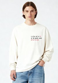 Chemise crème à manches longues avec un col rond. Présente un texte imprimé en noir et rouge sur le devant. Portée avec un jean bleu clair.