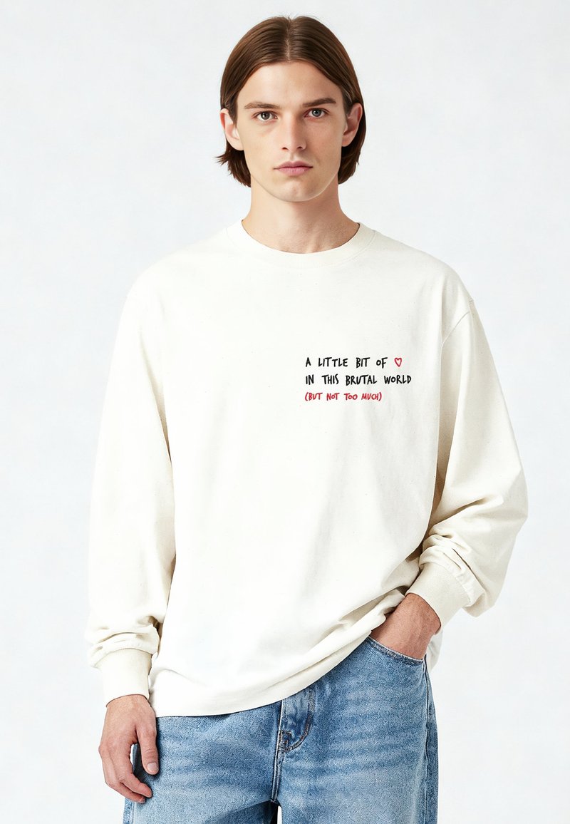 Chemise crème à manches longues avec un col rond. Présente un texte imprimé en noir et rouge sur le devant. Portée avec un jean bleu clair.