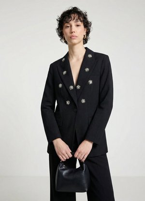 Femme aux cheveux courts et bouclés porte un blazer noir avec des broches décoratives étoiles argentées, un pantalon noir et tient un petit sac à main noir.