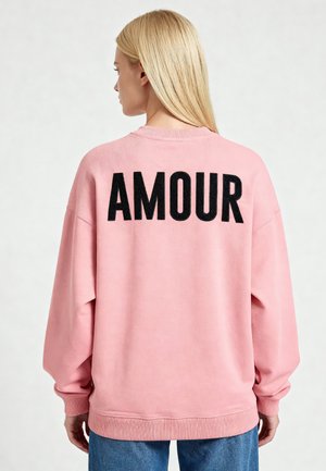 Femme blonde aux cheveux lisses portant un sweat-shirt rose avec « AMOUR » en lettres noires épaisses dans le dos, associé à un jean bleu.