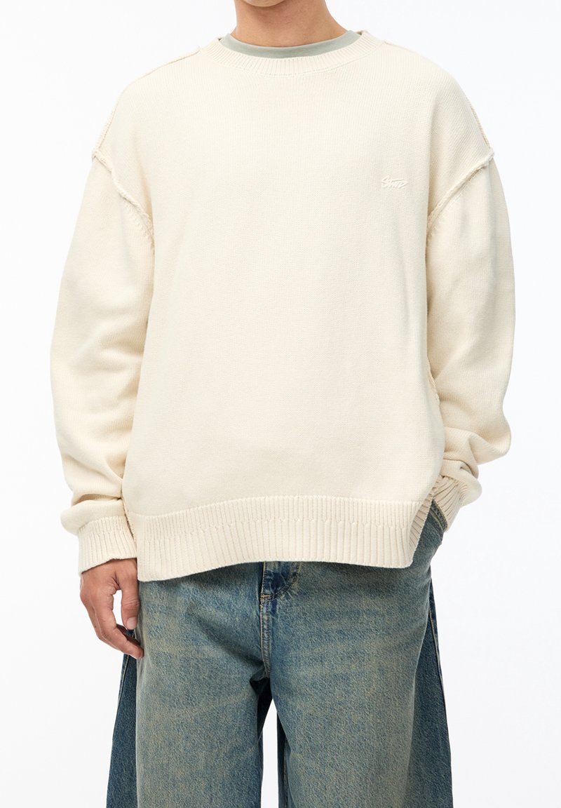 Homme portant un pull crème oversize et un jean bleu ample, avec une main dans la poche, montré du cou aux hanches sur fond blanc.