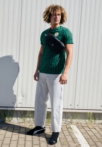 Grün gemustertes Kurzarmhemd, lockere helle Jeans, schwarze Bauchtasche, schwarze Sportschuhe mit weißen Akzenten vor einem grauen Hintergrund.