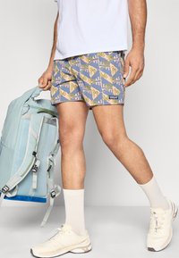 Μαγιό σορτς με μοτίβο φοίνικα σε μπλε, ροζ και πράσινο χρώμα· ελαφρύ ύφασμα· συνδυασμένο με ανοιχτό μπλε σακίδιο και εκρού αθλητικά παπούτσια.