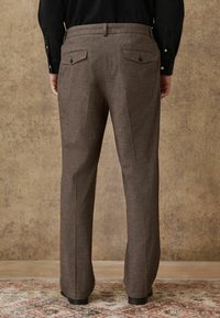 Brown, textured trousers with a straight cut. Features two back pockets with buttons and a waistband. Surface has a subtle pattern.