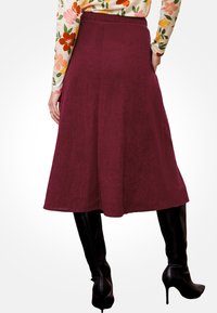 Mujer vistiendo una falda midi A-line burdeos, una blusa de manga larga con estampado floral y botas de tacón alto negras hasta la rodilla, de pie contra un fondo blanco.