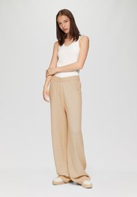 Beige wijde broek met een gestructureerde afwerking en een geribde, elastische tailleband, gecombineerd met een witte geribde tanktop. Eenvoudig, casual ontwerp.