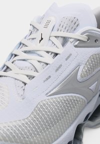 Κοντινό πλάνο αθλητικού παπουτσιού σε λευκό και γκρι, με ύφασμα από δίχτυ, συνθετικές επενδύσεις, λευκά κορδόνια και ασημένιο λογότυπο του εμπορικού σήματος στο πλάι.