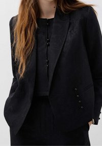 Mustavilla tekstuurilla oleva bleiseri, jossa on kauluspäällyste, viisi mustaa napin korostusta ja yksi rintatasku. Pukeutunut yhteensopivan mustan paidan päälle.