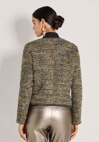 Frau mit dunklem Haar, das zu einem Dutt gebunden ist, trägt goldene geometrische Ohrringe, eine strukturiertes mehrfarbiges Strickjacke und metallisch silberne Hosen, von hinten blickend.