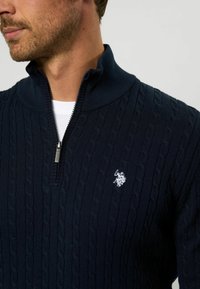 Jūras spēku zip-up džemperis ar teksturētu kabeli adījumu un baltu emblēmu akcentu uz krūšu. Augsts apkakle ar rāvējslēdzēja detaļu.