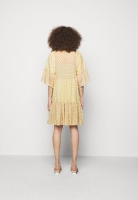 Kremowa sukienka o wzorzystej fakturze, z rozszerzanymi rękawami i warstwową spódnicą. Z tyłu widoczny jest otwór w kształcie łezki przy dekolcie.