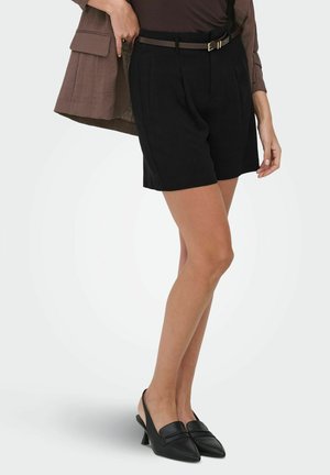 Vrouw draagt zwarte, hooggesneden, op maat gemaakte shorts met riem, een bruin ingestopt shirt, een bruine blazer en zwarte slingback kitten heels.