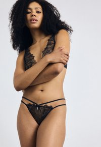 Ensemble de lingerie noire en dentelle florale transparente, avec un design en V profond et des bretelles fines. Détail avec un motif floral délicat et du matériel minimaliste.