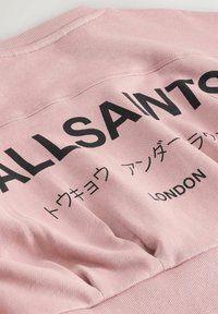Ružová mikina s čiernym potlačeným textom, vrátane "ALLSAINTS" a "LONDON", s jemnou, textúrovanou látkou a rebrovaným lemom.