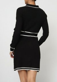 Robe noire côtelée à manches longues, dotée d'un col rayé blanc et d'une ceinture. L'ourlet présente des rayures blanches pour une touche de détail supplémentaire.