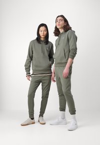 Olivgrüne Sweatshirts aus Baumwollmix mit Vorderaschen, kombiniert mit passenden, schmal geschnittenen Jogginghosen. Akzenten in Weiß und Beige bei den Sneakers.