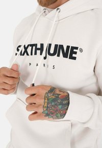 Hvit hettegenser med svart "SIXTHJUNE PARIS"-logo. Har snor, ribbestrikkede mansjetter og en kengurulomme foran. Detaljerte tatoveringer synlige på hånden.