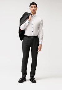 Camicia bianca con bottoni neri, abbinata a pantaloni grigi scuri su misura. Giacca nera appoggiata sulla spalla, cintura e scarpe nere.
