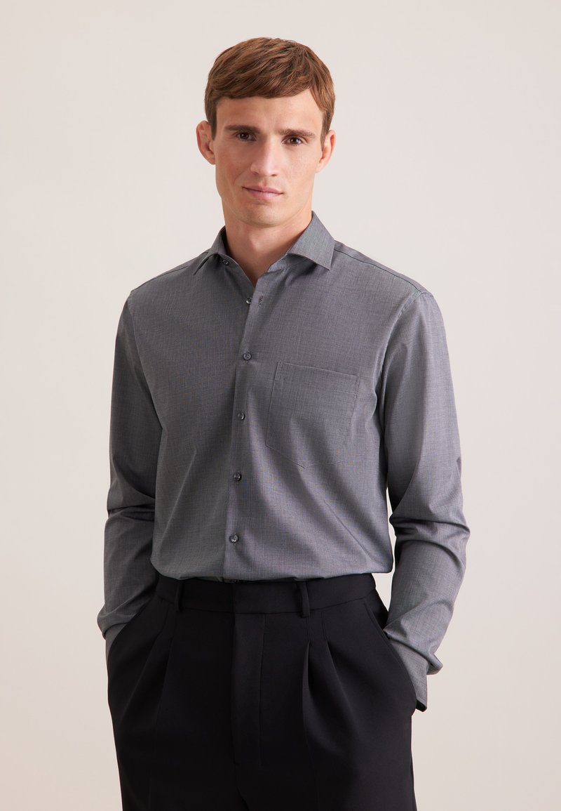 Homme portant une chemise grise à manches longues boutonnée, rentrée dans un pantalon noir plissé, debout les mains dans les poches sur un fond uni.