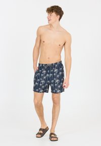 Marineblaue Schwimmshorts mit einem Palmenmuster, elastischem Bund, verstellbarem Kordelzug und seitlichen Taschen. Getragen mit braunen Sandalen.