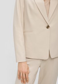 Beige blazer met een gestructureerde stof, enkele knoopsluiting en een subtiele zakdetail. Te combineren met beige pantalon.