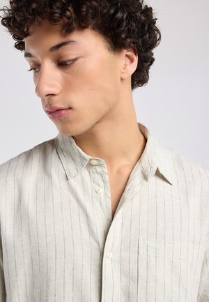 Jonge man met krullend haar kijkt naar beneden, draagt een lichtbeige dun gestreept overhemd met een borstzak.