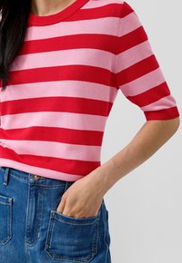 Persoon die een rood en roze gestreept T-shirt met korte mouwen draagt, met de hand in de voorspoort van een blauwe spijkerbroek, tegen een effen achtergrond.