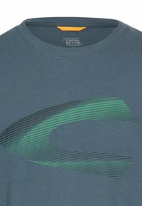 Blå crew-neck t-shirt med et grønt og sort abstrakt buet stribedesign på brystet og et lille Camel Active-logo inde i kraven.