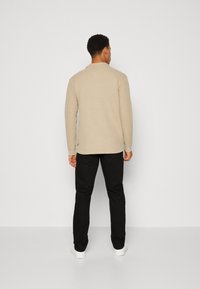 Un pull en tricot beige avec une finition texturée, porté avec un pantalon noir et des baskets blanches, vu de dos sur un fond uni.