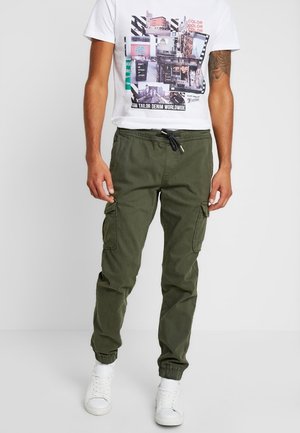 Homme portant un t-shirt blanc à motif graphique et un pantalon cargo vert olive avec taille à cordon et des baskets blanches, debout devant un fond uni.