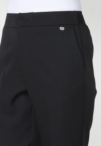 Pantalons noirs ajustés avec une texture lisse, des plis subtils et un petit logo métallique sur la taille.