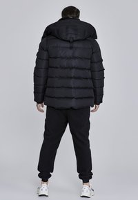 Sort polstret jakke med hætte, der har lodret quiltede sektioner og en robust lynlås. Sammen med sorte sweatpants og hvide sneakers.