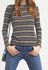 Schwarz-weiß gestreiftes Langarmoberteil mit goldenen Ketten- und gepunkteten Mustern, aus glattem Stoff gefertigt, kombiniert mit blauen Jeans.
