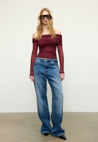 Weinrotes schulterfreies Oberteil mit gerafften Ärmeln, kombiniert mit blauen Jeans mit weitem Bein. Schwarze spitze High Heels und übergroße Sonnenbrillen.