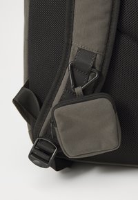 Sac à dos gris avec un tissu texturé, présentant un panneau en maille, des bretelles ajustables et une petite poche zippée attachée.