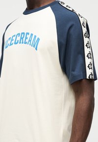 Κρεμ και ναυτικό μπλούζα raglan με το κείμενο "ICECREAM" σε μπλε χρώμα μπροστά, με διακοσμητική ταινία με σχέδια κατά μήκος των ώμων. Μαλακό βαμβακερό ύφασμα.