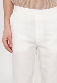 Pantalons en lin blancs avec une coupe décontractée et une taille élastique. Le tissu présente une texture lisse et un éclat subtil.