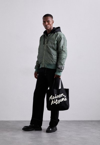 Πράσινο μπουφάν με φερμουάρ μπροστά, ριχτές μανσέτες και λεπτομερείς τσέπες. Μαύρη τσάντα ώμου με λευκό κείμενο "Maison Kitsuné". Μαύρα παντελόνια και παπούτσια.