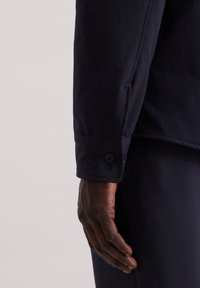 Granatowa marynarka zapinana na guziki o gładkiej fakturze. Widoczne detale mankietu z dwoma guzikami, dopasowany krój z eleganckim designem, z wysmakowanymi szwami.